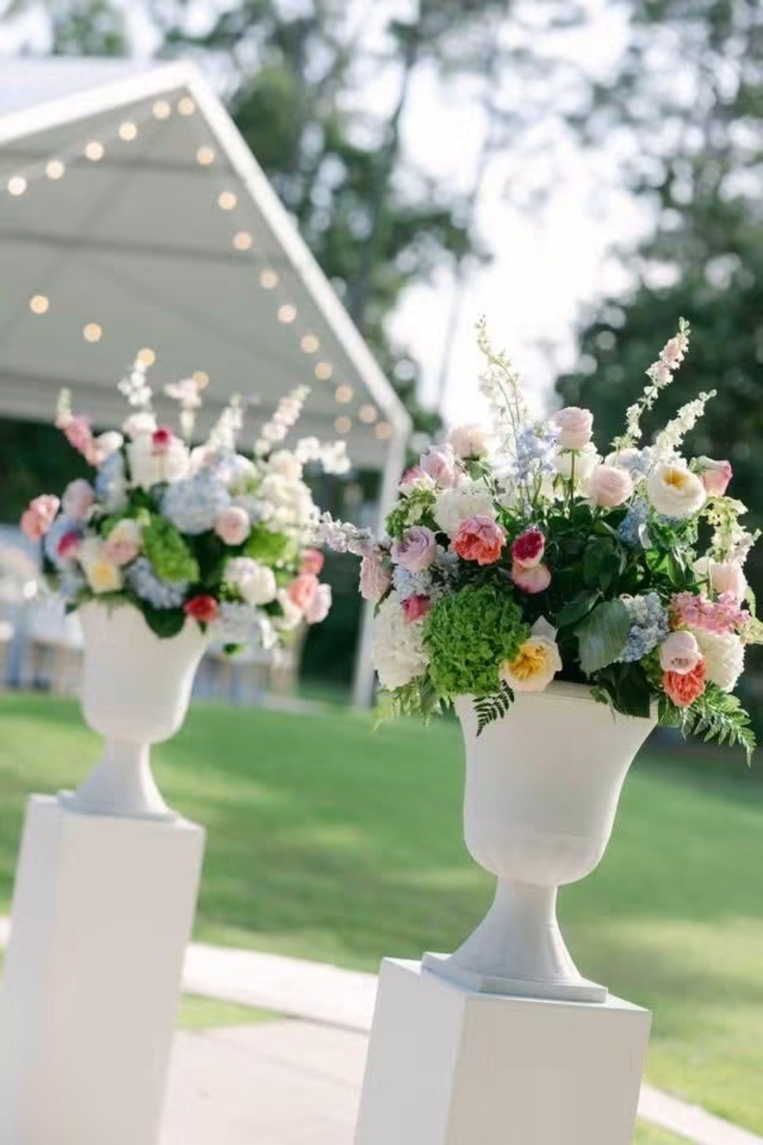 White urns of pastel florals on pedestals on a lawn